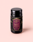 A tea canister labeled “Haunted Hibiscus: Tart Berry Herbal Blend with Warm Spices” features gold and purple decoration with botanical and mystical elements. It sits on a light pink background, and the label describes this caffeine-free blend as inspired by haunted hibiscus notes and vibrant hibiscus tea undertones.