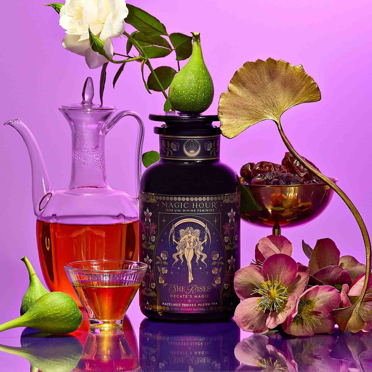 A canister of Hecate’s Magic: Hazelnut Toffee Puerh Tea for Calm &amp; Digestion surrounded by a glass carafe, a small bowl of dates, fresh figs, and pink flowers against a purple background.
