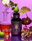 A canister of Hecate’s Magic: Hazelnut Toffee Puerh Tea for Calm & Digestion surrounded by a glass carafe, a small bowl of dates, fresh figs, and pink flowers against a purple background.