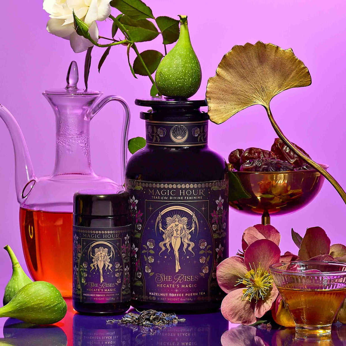 A table with the Monthly Magic Tea Journey Subscription container, a small jar, figs, dates, a red carafe, flowers, a tea glass, and a dried lotus leaf on a purple background.