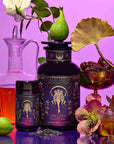 A table with the Monthly Magic Tea Journey Subscription container, a small jar, figs, dates, a red carafe, flowers, a tea glass, and a dried lotus leaf on a purple background.
