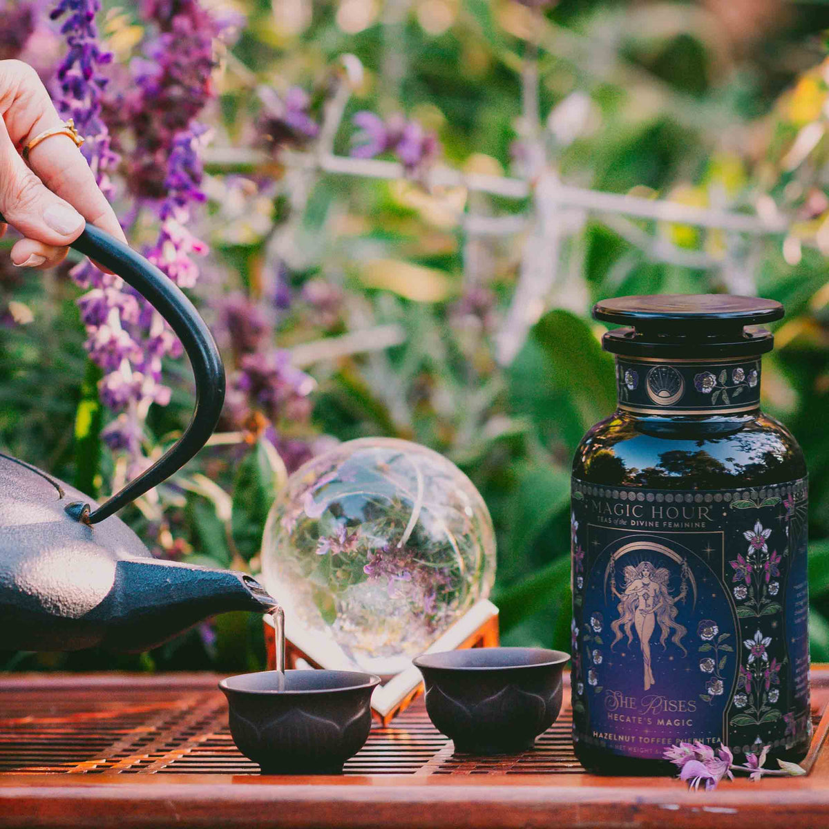 A hand pours tea from a black teapot into cups beside a dark floral jar labeled “Monthly Magic Tea Journey Subscription,” evoking ritual and Divine Feminine exploration amid purple flowers and lush greenery outdoors.