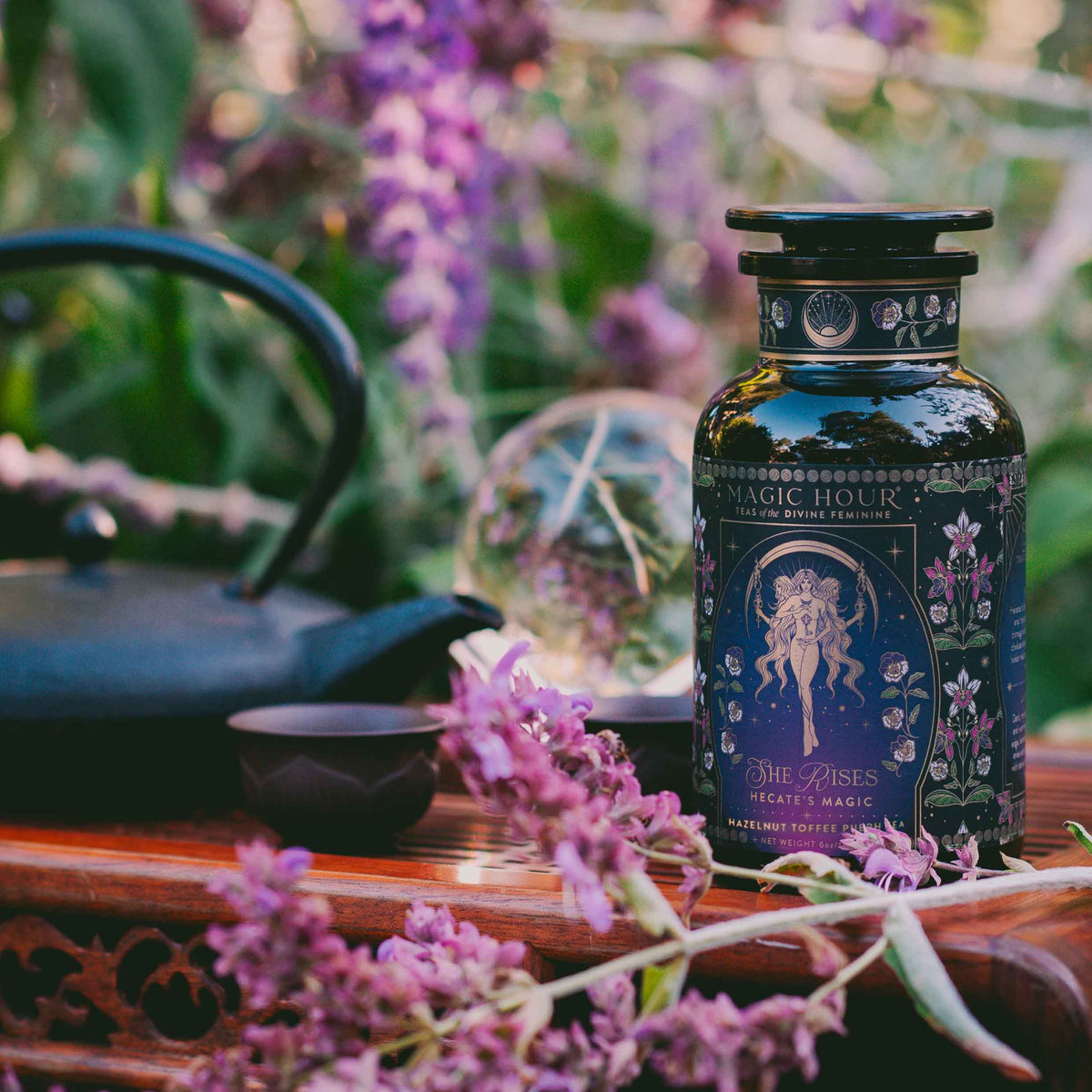 A decorative tea bottle from the Monthly Magic Tea Journey Subscription sits among purple flowers, a teapot, and a teacup outdoors—ideal for goddess ritual tea beneath lush greenery and sunlight.