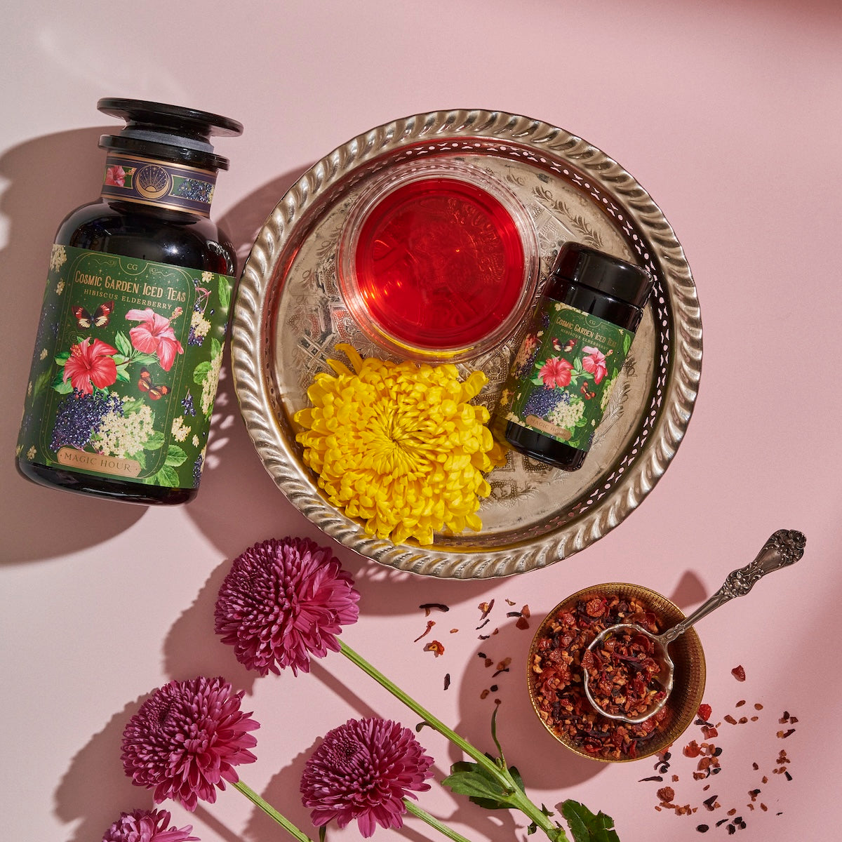 A jar and a smaller container of Magic Hour's Hibiscus Elderberry: Cosmic Garden Iced Tea are placed on a decorative silver tray, accompanied by a vibrant yellow flower and a clear glass of red liquid. The scene is bordered by purple flowers, and a spoon with dried loose leaf tea rests nearby.