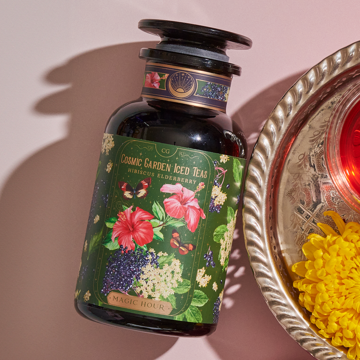 A dark glass bottle labeled "Hibiscus Elderberry: Cosmic Garden Iced Tea - Magic Hour" lies on a pink surface. The bottle features colorful illustrations of hibiscus flowers and butterflies, reminiscent of a Magic Hour Tea. Nearby, a silver tray with a red candle and yellow flowers is partially visible.