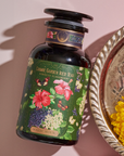 A dark glass bottle labeled "Hibiscus Elderberry: Cosmic Garden Iced Tea - Magic Hour" lies on a pink surface. The bottle features colorful illustrations of hibiscus flowers and butterflies, reminiscent of a Magic Hour Tea. Nearby, a silver tray with a red candle and yellow flowers is partially visible.