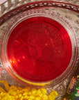 A round glass bowl filled with a bright red liquid is placed on an ornate silver tray. Beside the bowl, a yellow marigold flower and a colorful bottle with a floral design are visible, hinting at Hibiscus Elderberry: Cosmic Garden Iced Tea by Magic Hour. The setup appears decorative and festive.