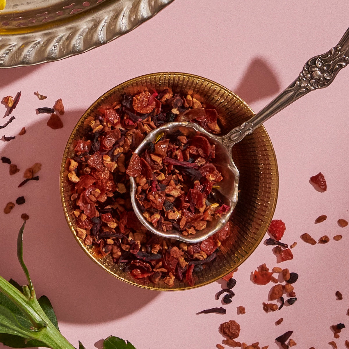 A close-up photo of a brass bowl filled with Hibiscus Elderberry: Cosmic Garden Iced Tea by Magic Hour, featuring a variety of red and brown pieces, with an ornate metal spoon resting inside. The organic tea is placed on a pink surface, and some of the loose leaf tea blend is scattered around. A green leaf is also partially visible.