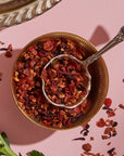 A close-up photo of a brass bowl filled with Hibiscus Elderberry: Cosmic Garden Iced Tea by Magic Hour, featuring a variety of red and brown pieces, with an ornate metal spoon resting inside. The organic tea is placed on a pink surface, and some of the loose leaf tea blend is scattered around. A green leaf is also partially visible.