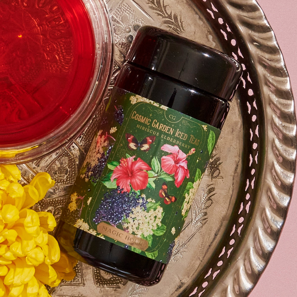 A tin labeled &quot;Hibiscus Elderberry: Cosmic Garden Iced Tea&quot; with floral artwork sits on an ornate silver tray. To its left is a glass of red liquid, and to its right are yellow flowers. The background features a soft pink surface, perfectly showcasing the allure of Magic Hour.