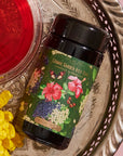 A tin labeled "Hibiscus Elderberry: Cosmic Garden Iced Tea" with floral artwork sits on an ornate silver tray. To its left is a glass of red liquid, and to its right are yellow flowers. The background features a soft pink surface, perfectly showcasing the allure of Magic Hour.