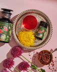 A jar and a smaller container of Magic Hour's Hibiscus Elderberry: Cosmic Garden Iced Tea are placed on a decorative silver tray, accompanied by a vibrant yellow flower and a clear glass of red liquid. The scene is bordered by purple flowers, and a spoon with dried loose leaf tea rests nearby.