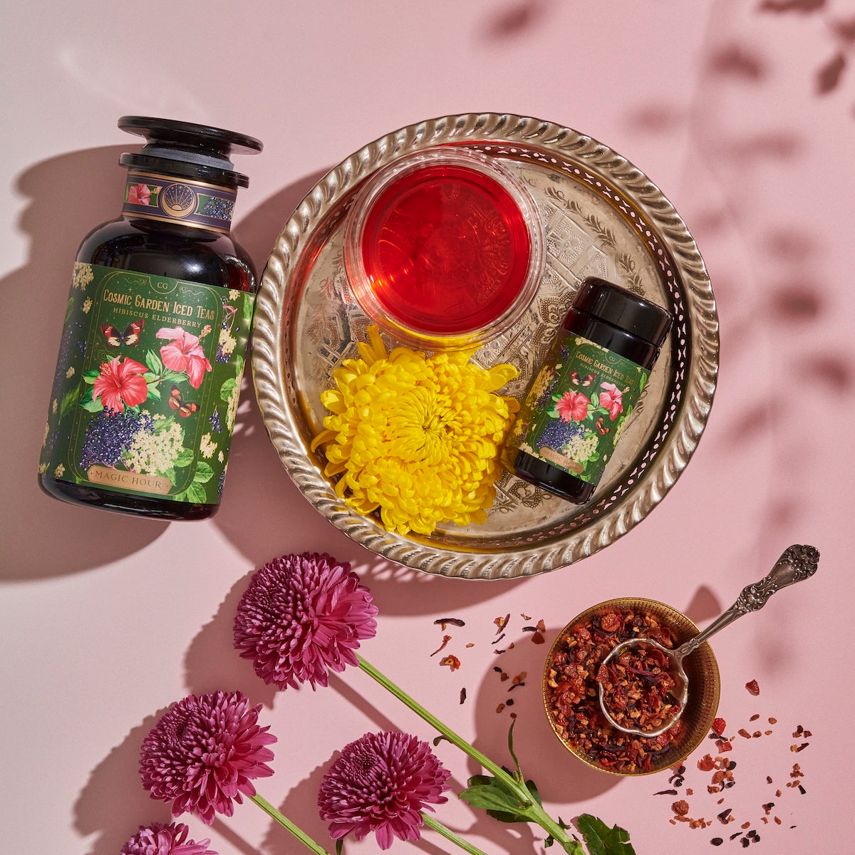 A floral-themed display features a bottle of Cosmic Garden Red Teas, a smaller bottle, a metal tray with a red liquid in a dish, and yellow chrysanthemum petals. A small bowl with a spoon contains herbal tea leaves. Purple flowers and scattered petals adorn the scene.