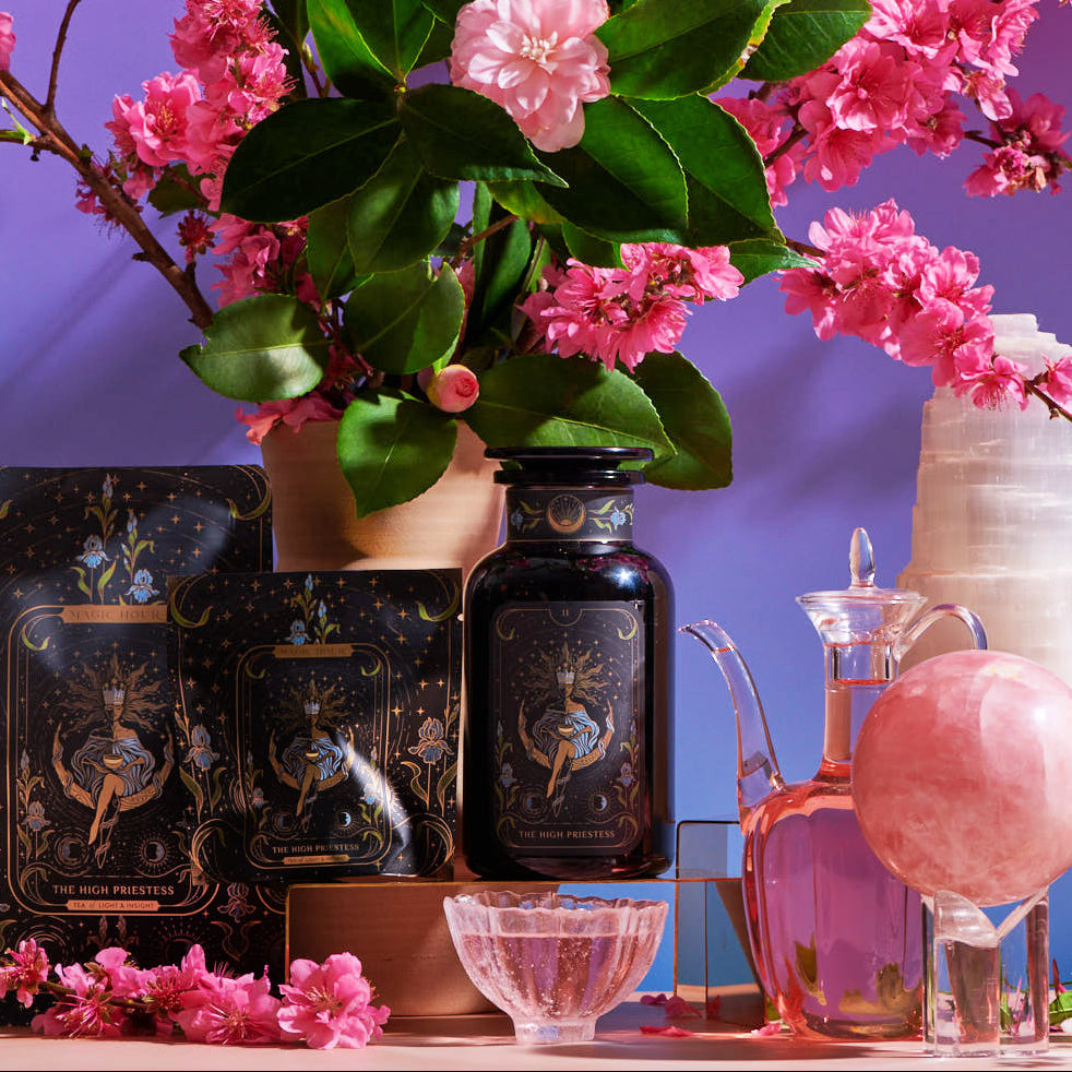 A decorative setup featuring The High Priestess Box skincare products with mystical-themed packaging, surrounded by pink cherry blossoms and green leaves. The display includes an ornate glass pitcher, a pink crystal sphere on a stand, a black crystal tower, and vintage glassware. A pastel background enhances the vibrant colors of The High Priestess Box.