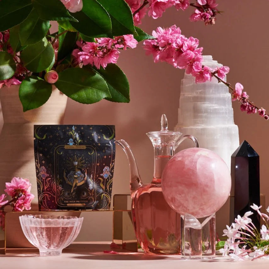 A decorative scene featuring pink blossoms, glass containers, and mystical objects. A tarot card evokes the 12 Tarot Tea Sampler Bundle experience, surrounded by a crystal orb, dark crystal tower, and tall white mineral formation. Pink flowers add vibrancy.