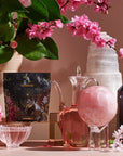 A decorative scene featuring pink blossoms, glass containers, and mystical objects. A tarot card evokes the 12 Tarot Tea Sampler Bundle experience, surrounded by a crystal orb, dark crystal tower, and tall white mineral formation. Pink flowers add vibrancy.