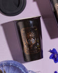 A black reusable cup with an artistic design labeled "The Queen of Cups - Tarot To Go!," featuring intricate, mystical imagery, perfect for savoring your favorite organic tea. The cup sits on a pink surface, accompanied by a matching lid, a blue flower, and part of a clear glass dish.