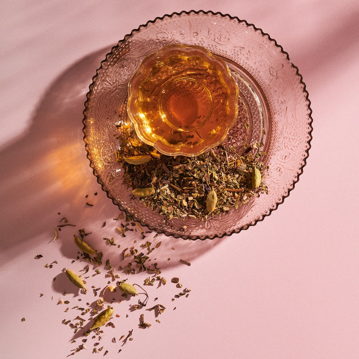 A glass teacup filled with The High Priestess: Wisdom Tea for Powerful Dreams & Illuminated Insights by Magic Hour sits on a decorative saucer with loose tea leaves and cardamom pods scattered around. The setup is on a pink surface, with the caffeine-free tea casting a warm amber hue against the saucer's intricate pattern.