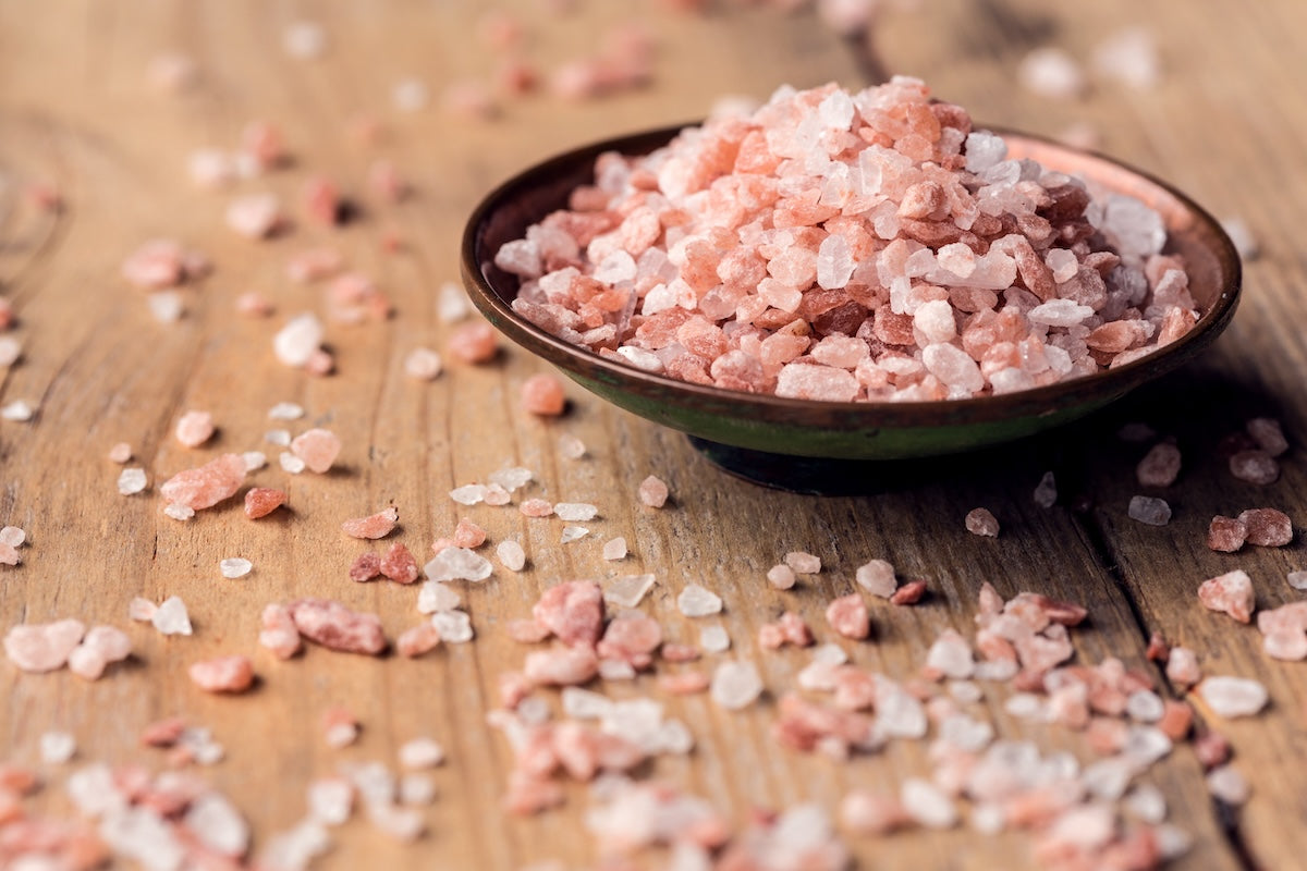 A small brown bowl filled with pink Himalayan salt crystals sits on a wooden surface, with additional salt crystals scattered around the bowl. The pink and white crystals contrast against the rustic wood background.