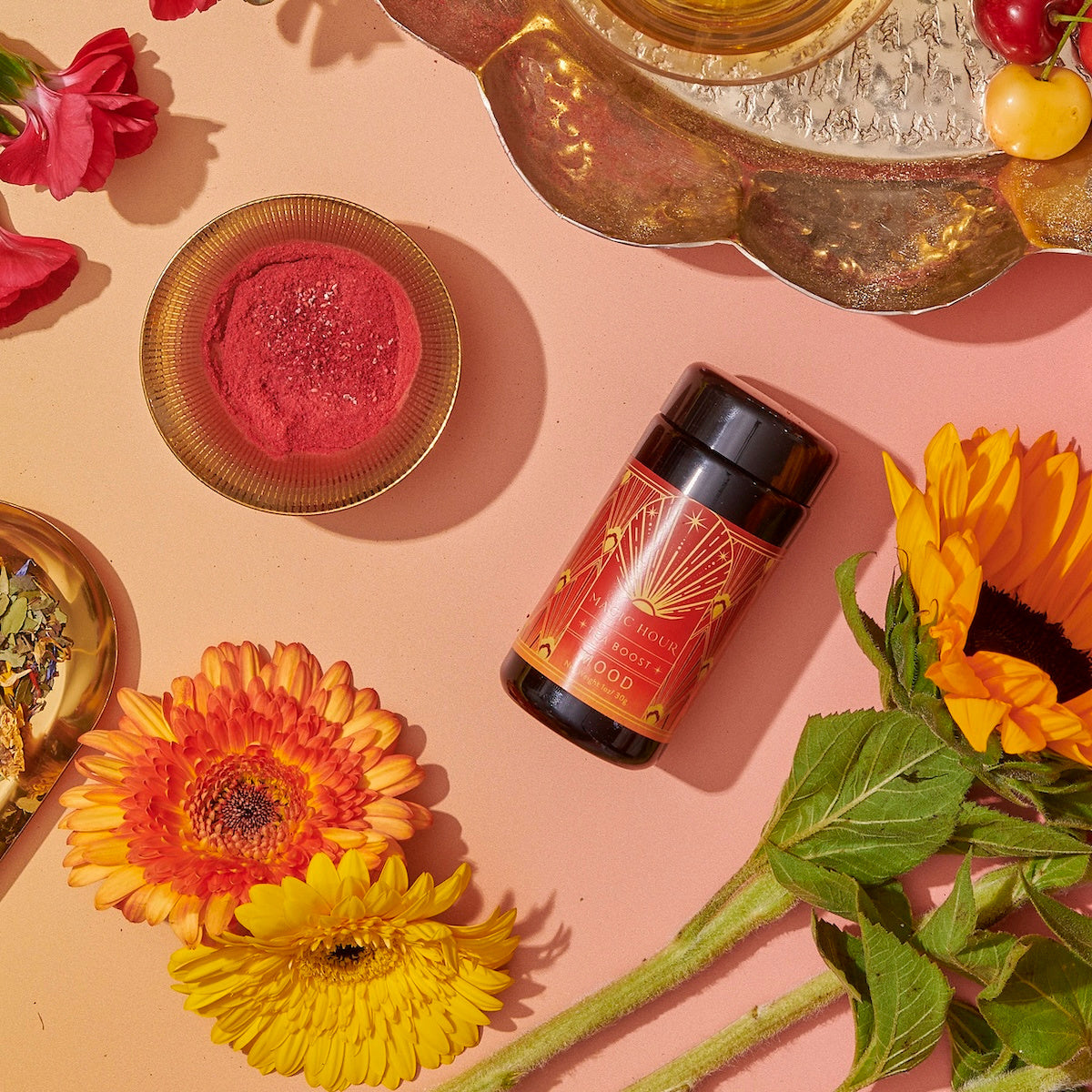 An arrangement features a black and red bottle of Hydrate Tea Boost with gold accents, a small dish of red powder, and a variety of flowers including sunflowers and orange gerbera daisies. A decorative tray with assorted items is partially visible in the background.