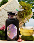 A jar of Perfumes of Provence: Lavender Bouquet Tea for Calm Moods and Beautiful Skin sits by a glass cup of tea, surrounded by moss, pink flowers, a fern, and a dragonfly decoration, with a large white stone behind.