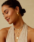 A woman with dark hair in a low bun smiles with her eyes closed, showcasing gold hoop earrings and multiple layered 18KT gold plate necklaces, including the stunning Inward Journey Pearl Mandala Necklace. She is wearing a light beige, sleeveless top and stands against a neutral background.