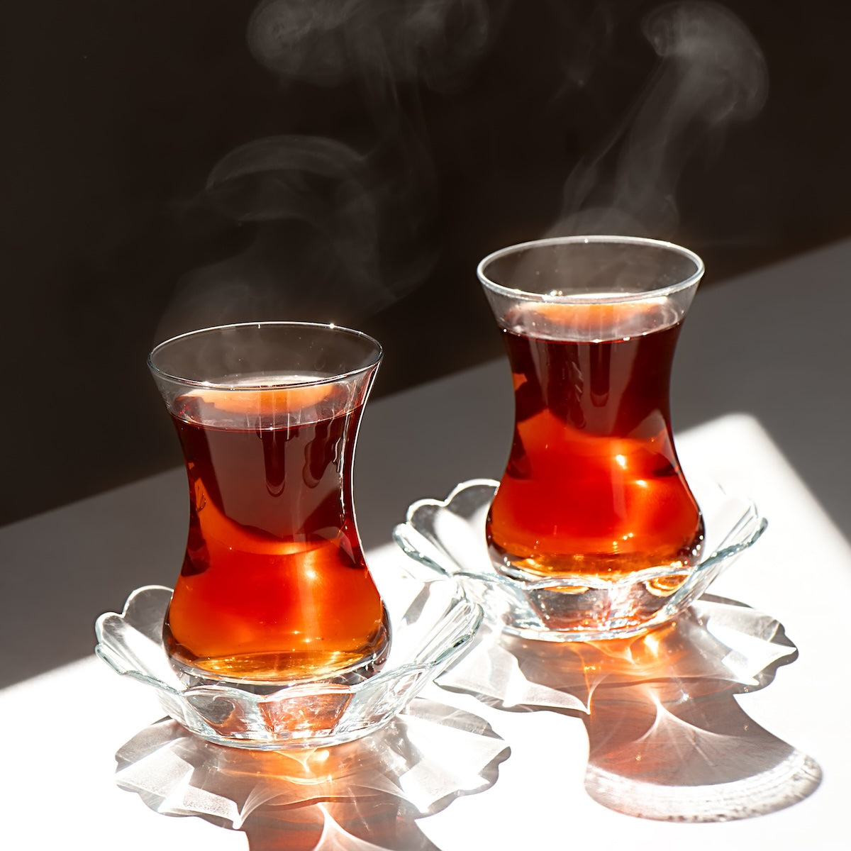 Two Turkish tea glasses filled with hot Organic Iyerpadi Estate Black Tea rest on glass saucers, steam swirling upward as sunlight highlights reflections and shadows across the white surface beneath them.