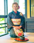 A woman kneeling on a tatami mat, adorned in a traditional black kimono with floral patterns and a colorful obi, smiles serenely in a Japanese-style room with sliding doors and views of a garden. In front of her lies the Tea School 2024-2025 Full Course Bundle by Magic Hour, displayed on a red lacquer tray with stacked bowls, echoing the traditions of the Japanese Tea School.