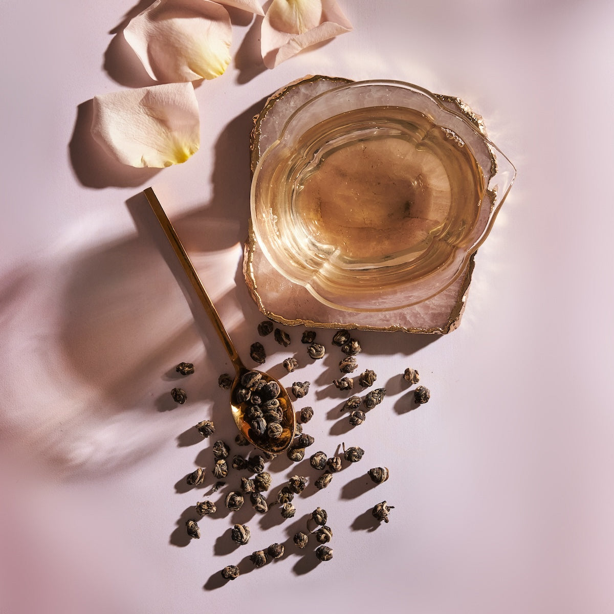 A delicate translucent tea cup with gold edges sits on a pink surface. Beside it, a gold spoon holds Jasmine Pinnacle Pearls Green Tea from Magic Hour, with more pearls scattered around. Light pink rose petals and jasmine flowers are arranged nearby, adding a soft, elegant touch to the serene scene.