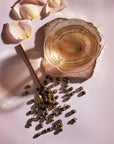 A delicate translucent tea cup with gold edges sits on a pink surface. Beside it, a gold spoon holds Jasmine Pinnacle Pearls Green Tea from Magic Hour, with more pearls scattered around. Light pink rose petals and jasmine flowers are arranged nearby, adding a soft, elegant touch to the serene scene.