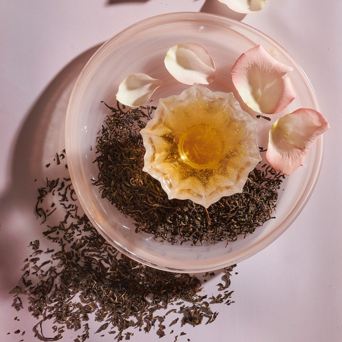A glass bowl filled with loose Magic Hour Jasmine Yin Hao Green Tea and a scoop of honey sits on a light pink surface. Rose petals are arranged around the bowl, creating an elegant and soothing composition. The setup is bathed in soft, natural light.