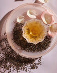 A glass bowl filled with loose Magic Hour Jasmine Yin Hao Green Tea and a scoop of honey sits on a light pink surface. Rose petals are arranged around the bowl, creating an elegant and soothing composition. The setup is bathed in soft, natural light.