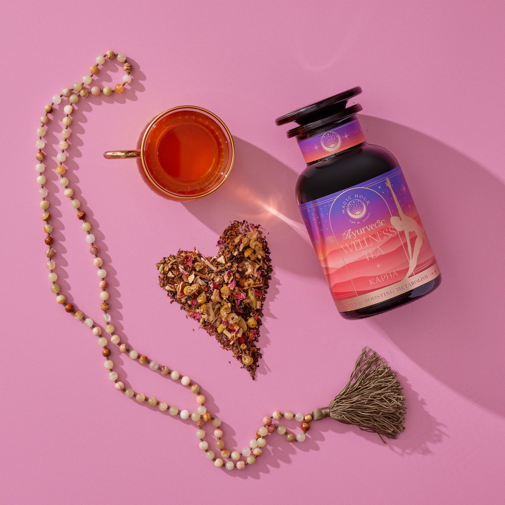 A bottle of Kapha Herbal capsules, a cup of tea, a heart-shaped pile of loose tea leaves and herbs, and a beaded tassel necklace are arranged on a pink surface.