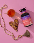 A bottle of Kapha Herbal capsules, a cup of tea, a heart-shaped pile of loose tea leaves and herbs, and a beaded tassel necklace are arranged on a pink surface.