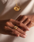 Close-up of a person's hands wearing the Keeper of Positivity Eye Blue Topaz Ring, featuring an eye-shaped design with a blue topaz at its center. The person's chest displays a matching gold necklace with a similar evil eye pendant. The background is softly lit, highlighting the jewelry and the person's light, neutral-colored clothing.