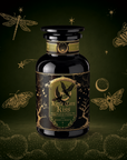 A dark glass bottle labeled “Kettle Corn Kiss: Sweet & Nutty Toasted Rice Green Tea with Caramel & Vanilla” sits on a green background adorned with gold dragonflies, moths, a crescent moon, stars, and foliage—evoking a whimsical, magical feel that perfectly captures the enchanting flavors of caramel and vanilla blended with the sweet nuttiness of toasted rice green tea inside.
