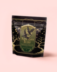 A black and gold pouch of Kettle Corn Kiss: Sweet & Nutty Toasted Rice Green Tea with Caramel & Vanilla, decorated with an owl, stars, and trees, sits upright against a light pink background.