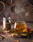 A steaming glass mug of Kettle Corn Kiss—a sweet & nutty toasted rice green tea with caramel & vanilla—rests on a wooden board by autumn leaves and a jar of loose tea, with rustic kitchen decor in the background.