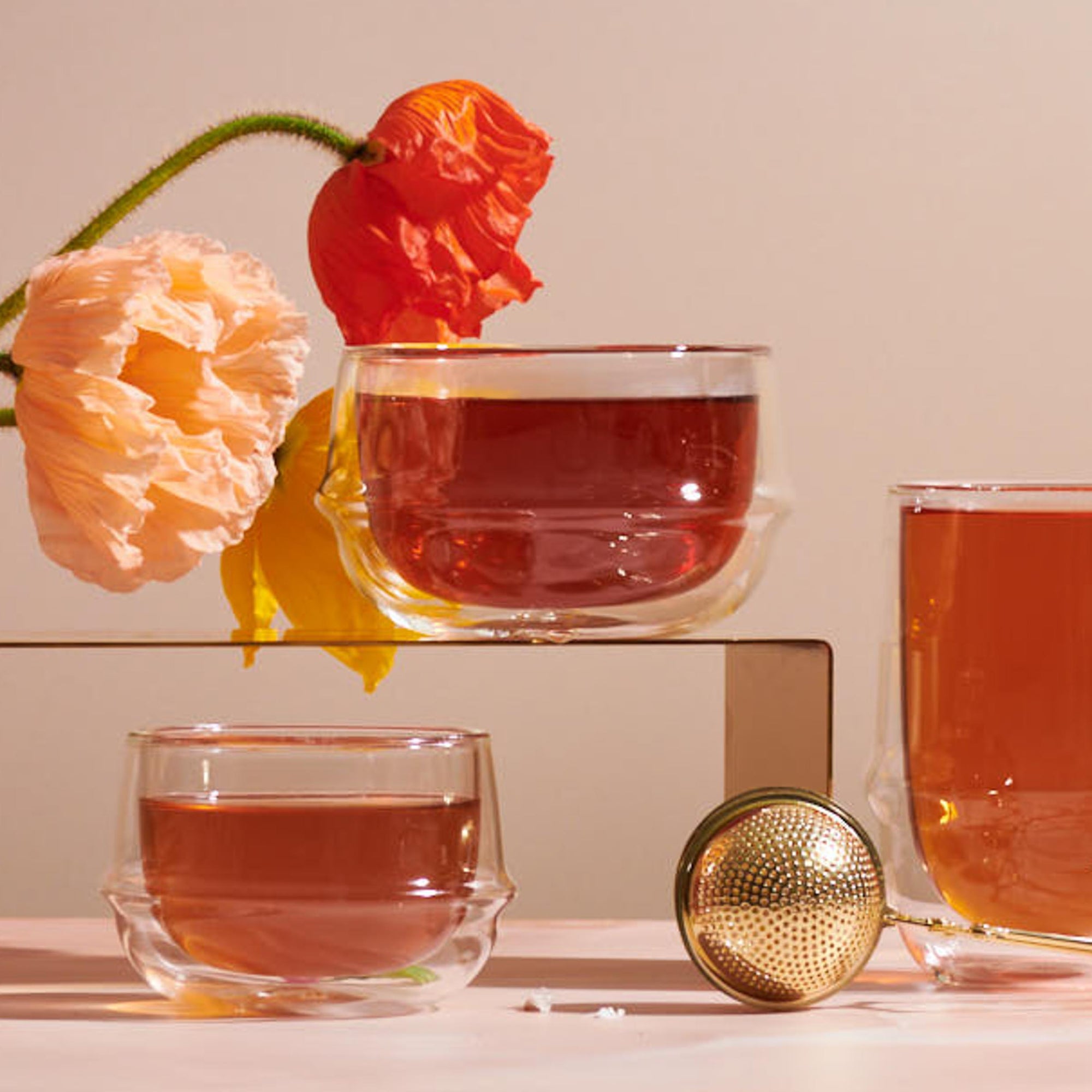 Two Kinto Double-Walled Teacups filled with tea sit beside a metal tea infuser. Two colorful poppy flowers hang over the scene, and a third glass of tea rests in the background—all arranged on a light pink surface.