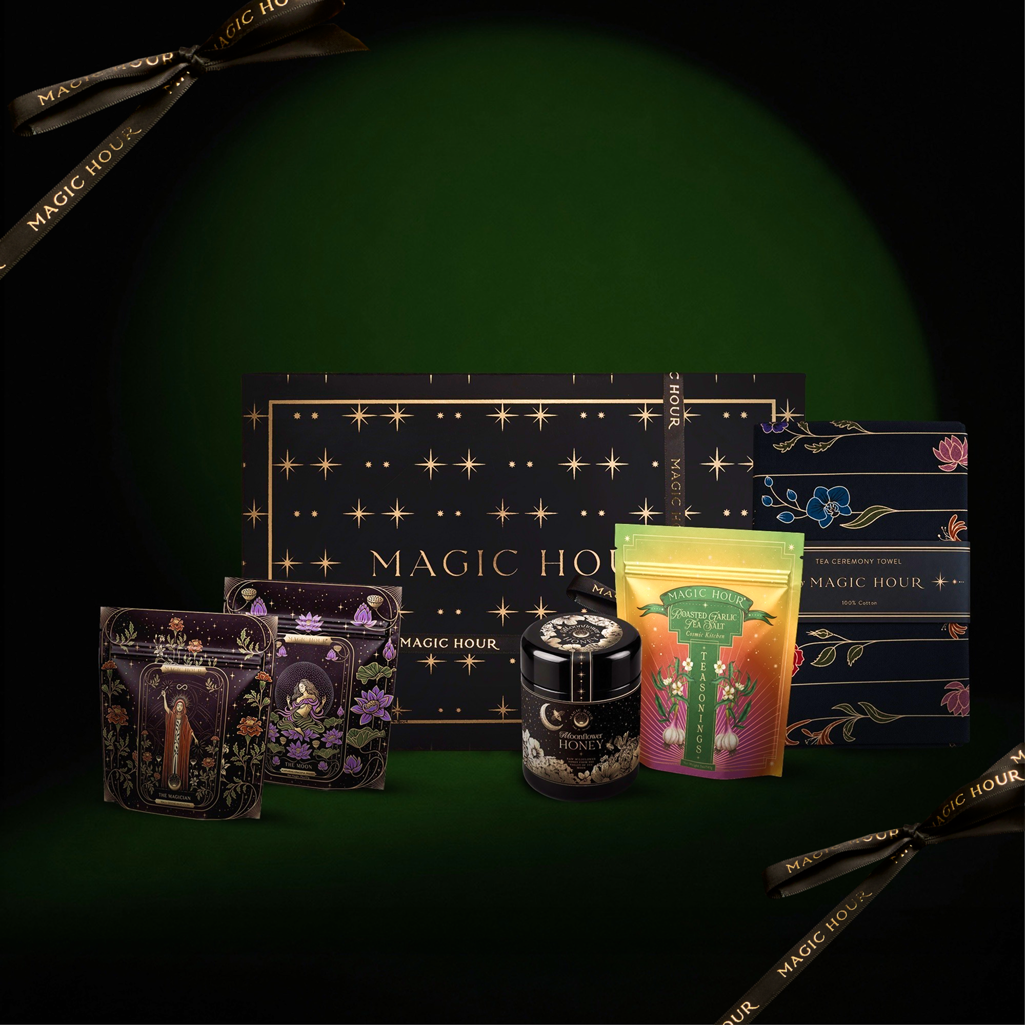 The Kitchen Witch Starter Box features a vibrant assortment of elegantly packaged tea products—including boxes, tins, and tea bags—adorned with “Magic Hour” branding set against a deep green backdrop with gold decorative accents. This enchanting collection captures the whimsical spirit of a kitchen witch and pairs perfectly with wildflower honey for an extra hint of sweetness.