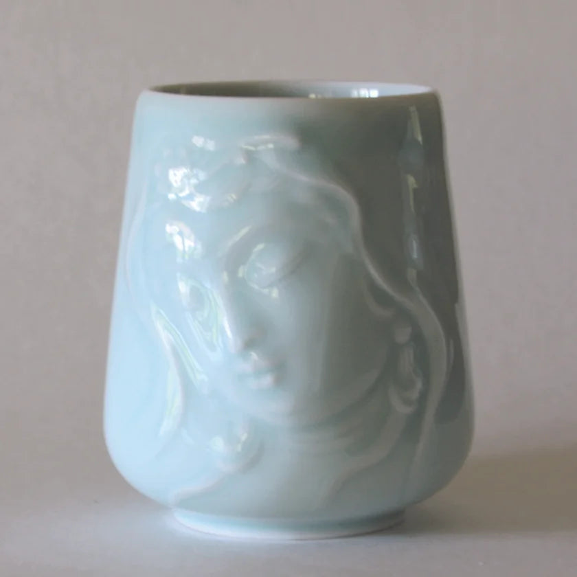 The Kwan Yin Face Mug by Eastern Elm is a celadon blue ceramic cup adorned with an embossed design of a serene face with closed eyes. It boasts a smooth, glossy finish and gently flares outward toward the base, perfectly capturing the peaceful essence of Kwan Yin.