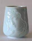 The Kwan Yin Face Mug by Eastern Elm is a celadon blue ceramic cup adorned with an embossed design of a serene face with closed eyes. It boasts a smooth, glossy finish and gently flares outward toward the base, perfectly capturing the peaceful essence of Kwan Yin.