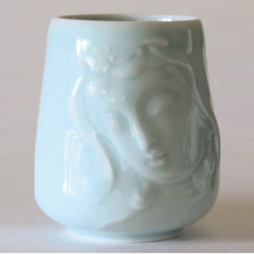 Introducing the Kwan Yin Face Mug by Eastern Elm, a celadon blue ceramic piece with a glossy finish. The mug showcases an embossed depiction of Kwan Yin's serene face with closed eyes and flowing hair, exuding an artistic and elegant charm against its plain off-white background.