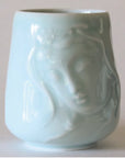 Introducing the Kwan Yin Face Mug by Eastern Elm, a celadon blue ceramic piece with a glossy finish. The mug showcases an embossed depiction of Kwan Yin's serene face with closed eyes and flowing hair, exuding an artistic and elegant charm against its plain off-white background.