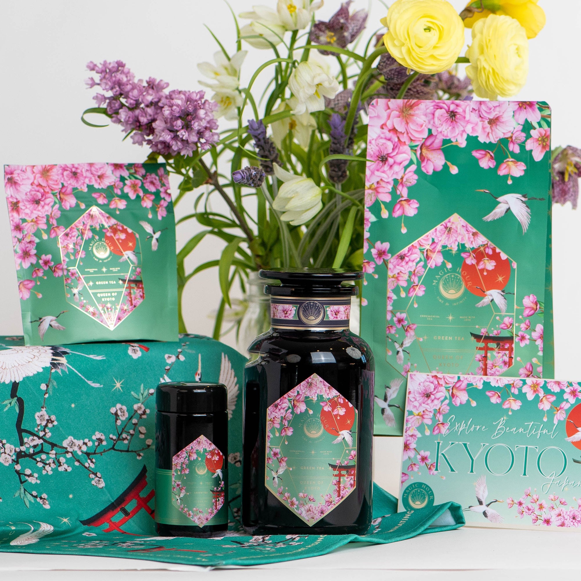 A display of Queen of Kyoto beauty products with Japanese Cherry Blossom and crane designs, set against colorful flowers. Packaging features a green and pink floral theme with "Kyoto" labeled on each item.