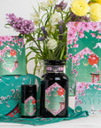 A display of Queen of Kyoto beauty products with Japanese Cherry Blossom and crane designs, set against colorful flowers. Packaging features a green and pink floral theme with "Kyoto" labeled on each item.
