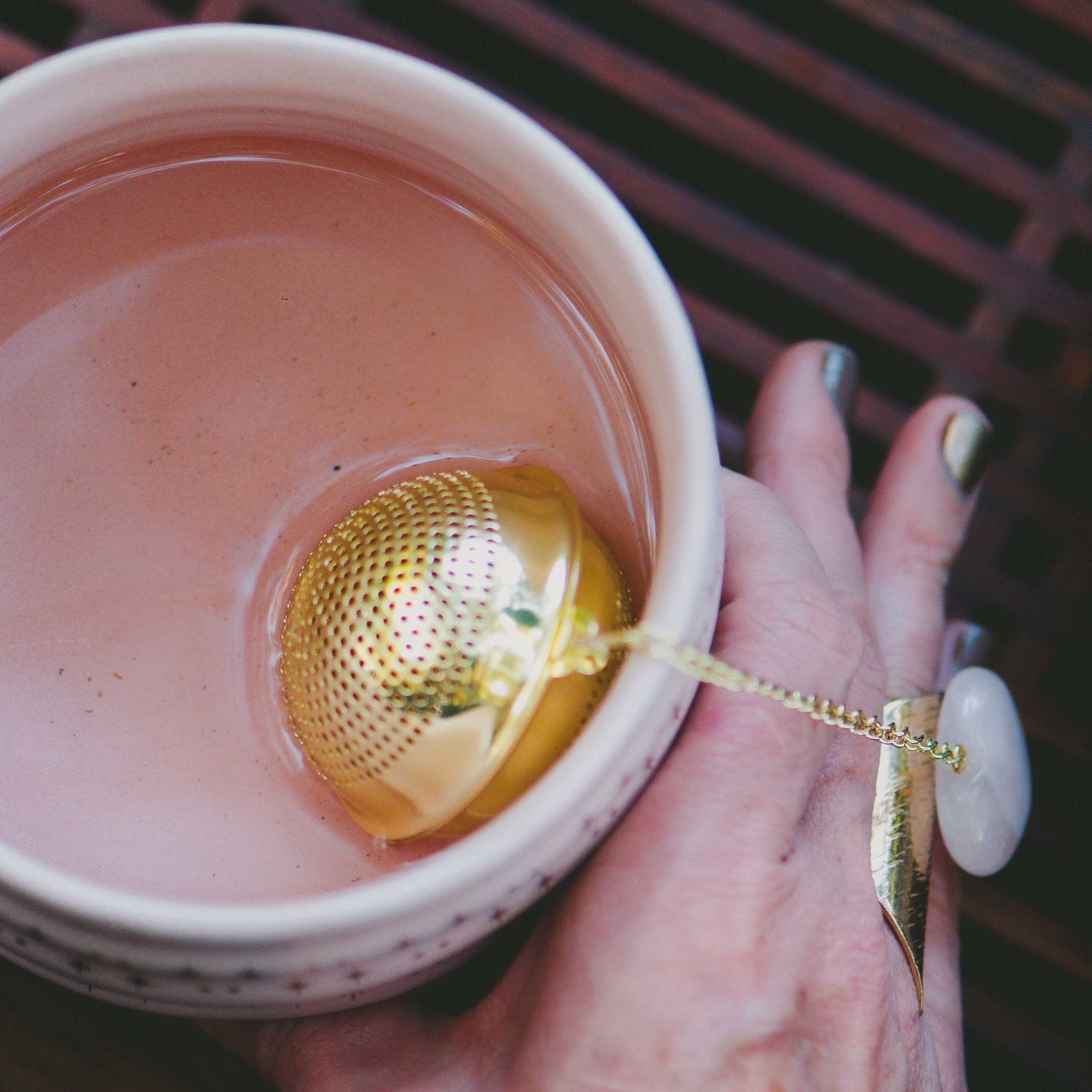 A hand with painted nails holds a pink cup of tea, featuring the Rose Quartz Heart Tea Strainer inside. The elegant scene is enhanced by a large white and gold ring worn by the person, adding sophistication to this peaceful tea ritual.
