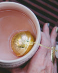 A hand with painted nails holds a pink cup of tea, featuring the Rose Quartz Heart Tea Strainer inside. The elegant scene is enhanced by a large white and gold ring worn by the person, adding sophistication to this peaceful tea ritual.