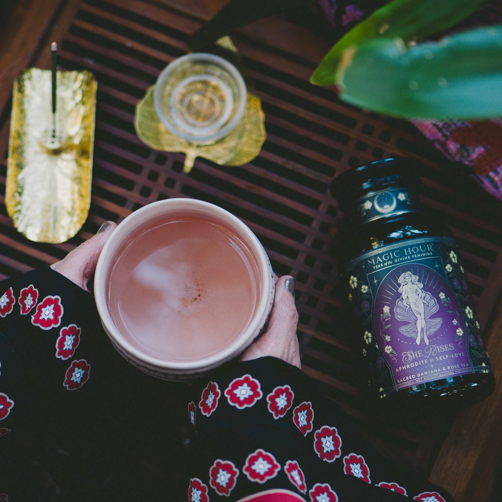 A person in a patterned sleeve holds a mug of tea above a wooden surface, near a jar of Aphrodite's Alchemy: Damiana & Rose White Tea, a gold incense holder with burning incense, and a glass of tea—evoking calming rituals.