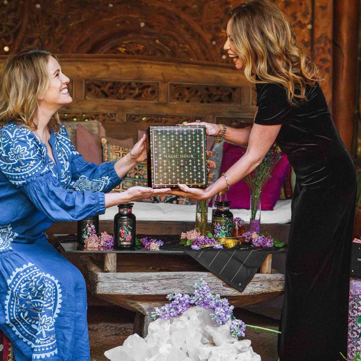 Two women smile as they exchange a Branded Gift Box, surrounded by flowers, tea jars, and crystals on a cozy, ornate wooden bench.